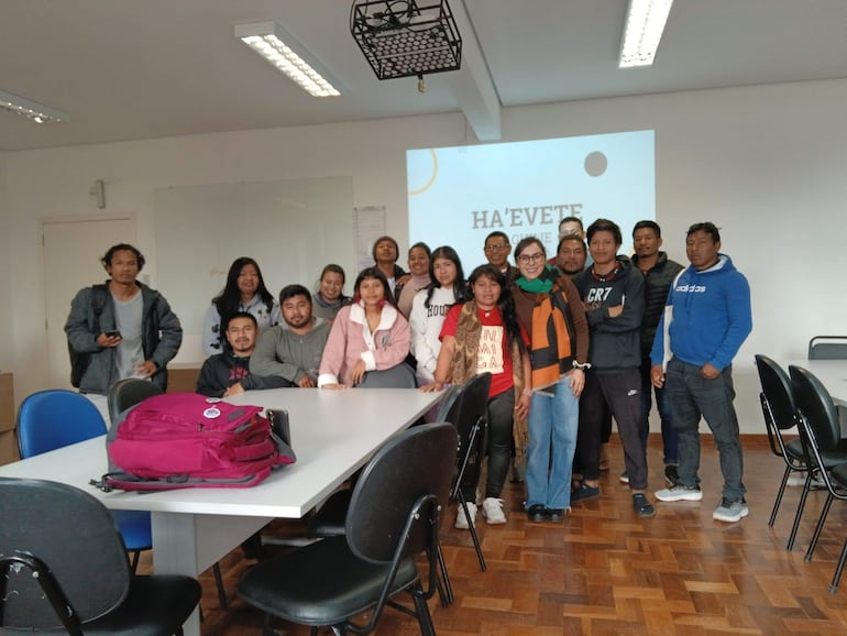 Grupo de jóvenes sonrientes en aula, algunos de pie, otros sentados, con mochila rosa sobre la mesa.