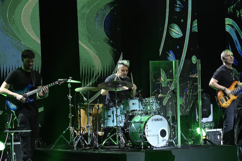 Tres músicos de Los Nocheros en el escenario, uno toca guitarra eléctrica azul, otro batería y el tercero bajo, rodeados de luces verdes.