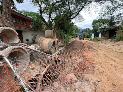 Escena de una obra en Santo Domingo con retroexcavadora en movimiento y tubos de concreto apilados al lado de un jardín.