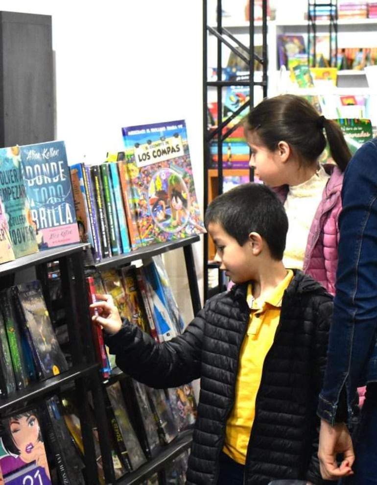 Dos niños en librería: uno sostiene la mano de otro mientras examina un libro entre estanterías coloridas y modernas.