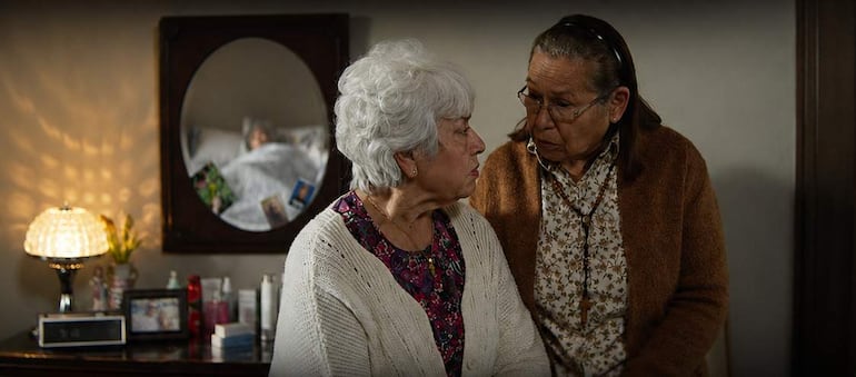 Dos mujeres mayores comparten una conversación en un entorno acogedor, con lámpara de mesa y productos de cuidado personal en la mesita.