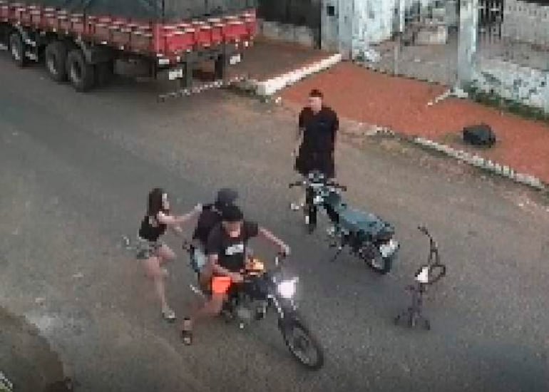 Mujer con cabello largo y camiseta ayuda a hombre con camiseta negra sobre motocicleta, observados por un tercero en ambiente urbano.