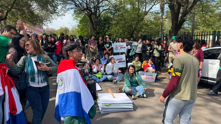 Jóvenes manifestantes sostienen carteles con mensajes de protesta, algunos aplauden, mientras otros escuchan con atención en un ambiente urbano.