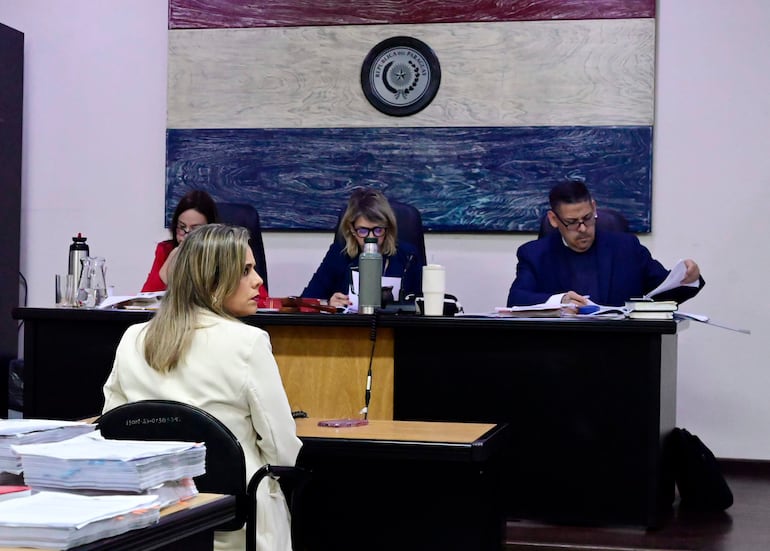 Kattya Mabel Gonzalez Villanueva, de espaldas, viste chaqueta blanca, en audiencia formal con documentos sobre la mesa y ambiente judicial.
