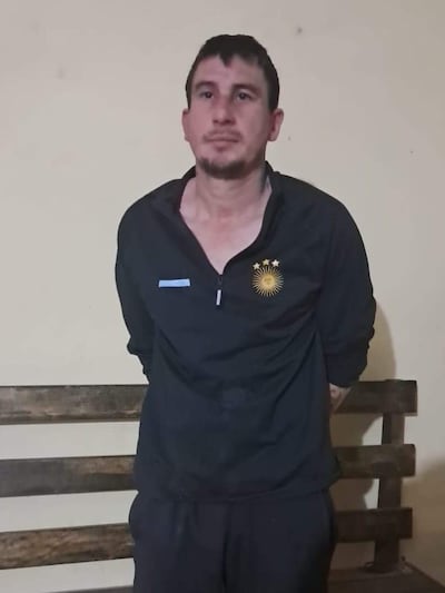 Hombre de pie, vestido con camiseta negra, expresión neutral, en una sala de detención con fondo claro y banco de madera.