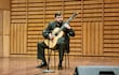 Javier Acosta Giangreco tocando la guitarra acústica en un auditorio con traje oscuro y fondo de madera iluminado.