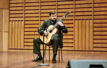 Javier Acosta Giangreco tocando la guitarra acústica en un auditorio con traje oscuro y fondo de madera iluminado.