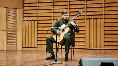 Javier Acosta Giangreco tocando la guitarra acústica en un auditorio con traje oscuro y fondo de madera iluminado.