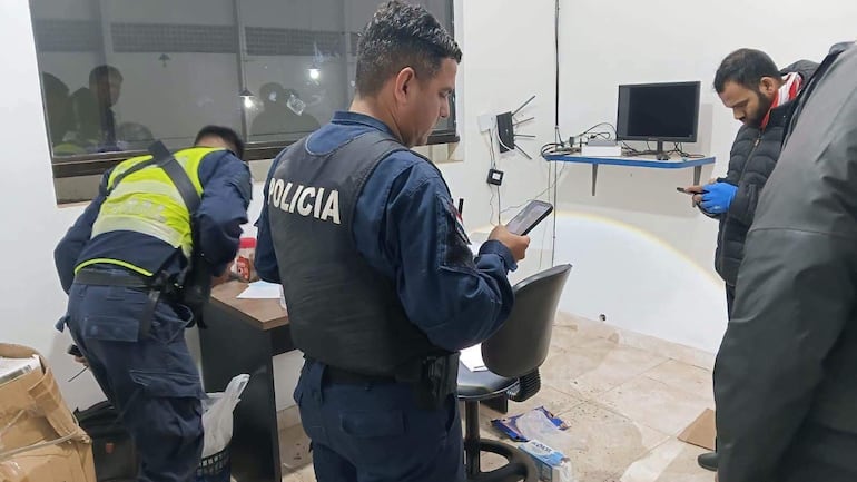 Dos policías en uniforme revisan documentos mientras otro hombre en abrigo negro utiliza su teléfono movil en un ambiente de oficina.