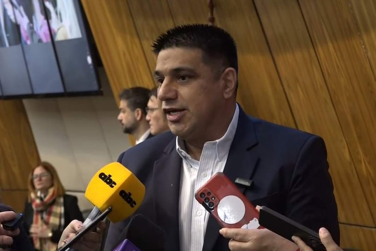Hugo Meza, con saco oscuro y camisa clara, responde a preguntas ante micrófonos. Varios periodistas lo observan atentamente.