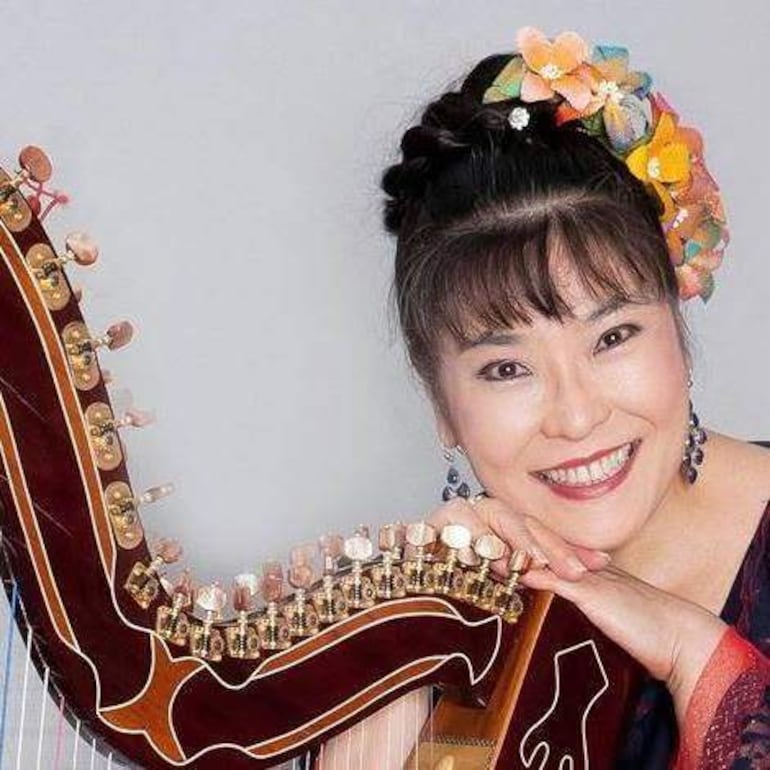 Lucía Shiomitsu sonríe de pie con un atuendo colorido y una harpa a su lado, en un fondo neutro.