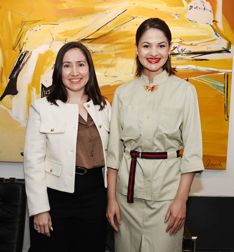 Dos mujeres sonrientes en atuendos formales posan junto al cónsul en un entorno elegante con obra de arte colorida de fondo.