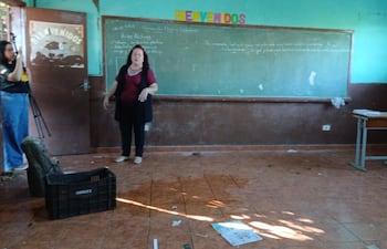 Mujer con blusa roja y chaleco negro habla en un aula desordenada con paredes verdes y marrones, letrero de 'BIENVENIDOS' visible.
