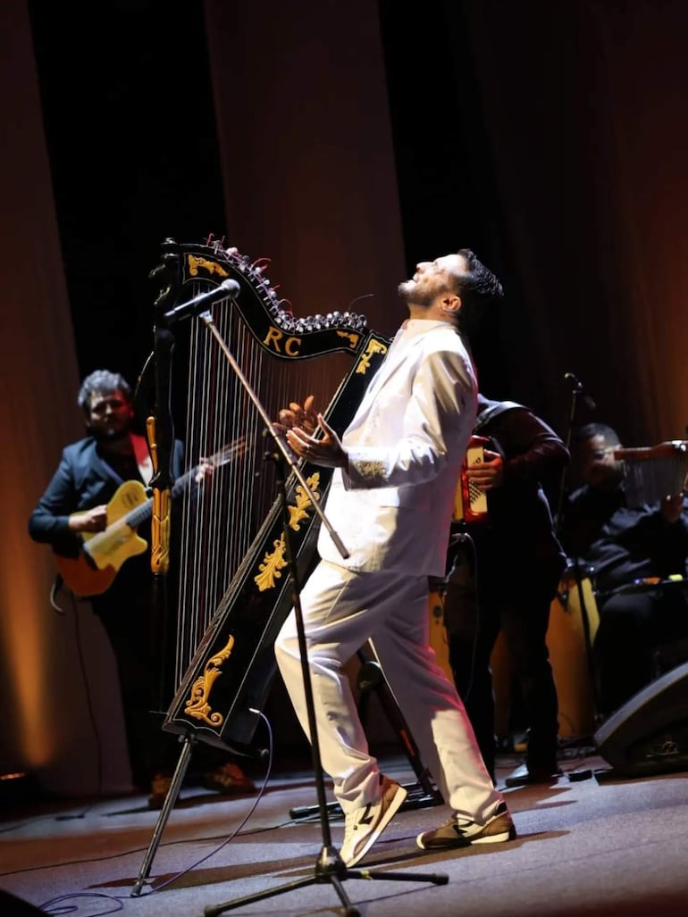 Músico con traje blanco tocando el arpa enérgicamente, rodeado de otros tres artistas en un escenario iluminado.