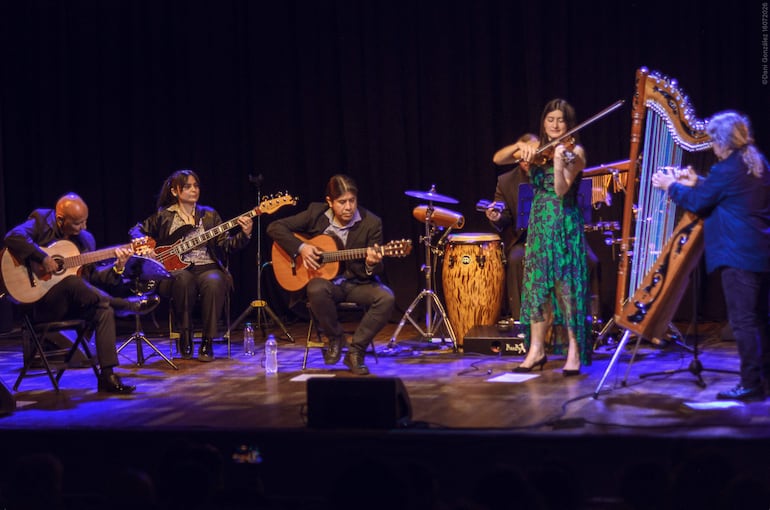 Músicos en el escenario iluminado por luces azules, una mujer con vestido verde toca el violín mientras otros instrumentistas los acompañan.