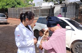 Profesional de salud en bata blanca interactúa con mujer mayor en abrigo rosa y niña en el exterior del hospital.