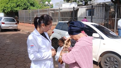 Profesional de salud en bata blanca interactúa con mujer mayor en abrigo rosa y niña en el exterior del hospital.