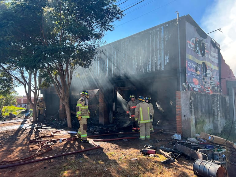 Cuatro bomberos en trajes de protección y cascos ante un edificio dañado, con humo y mangueras esparcidas a su alrededor.