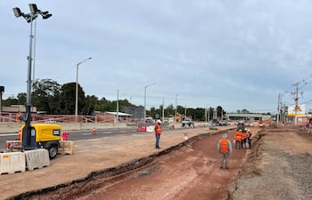 Seis trabajadores con chalecos naranjas y cascos en un sitio de construcción; maquinaria amarilla y conos de tráfico visibles.