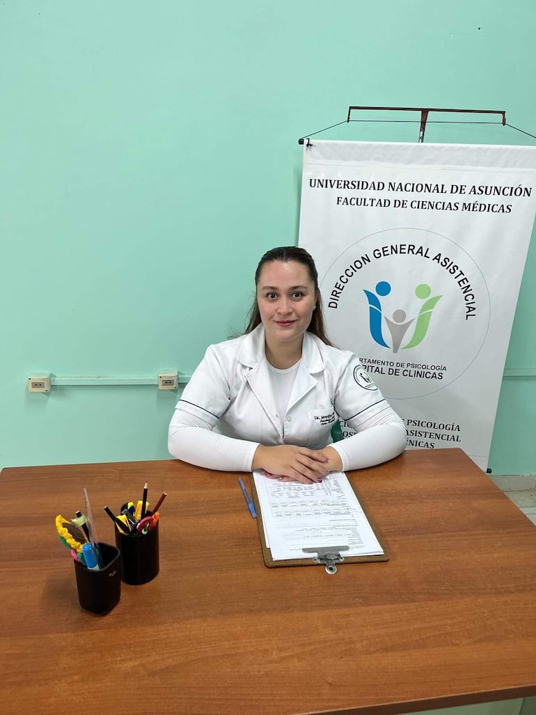 Lic. Jennifer Hidalgo Morel con uniforme médico blanco, sentada ante un documento, lista para hablar en un ambiente académico con banner institucional.