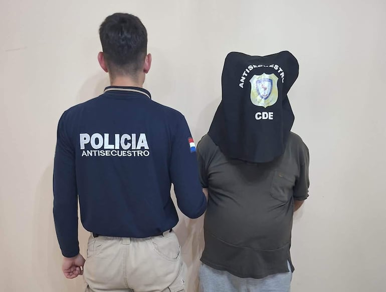 Policía antiextorsión en camiseta oscura supervisa a hombre cubierto con saco negro, en ambiente interior claro.