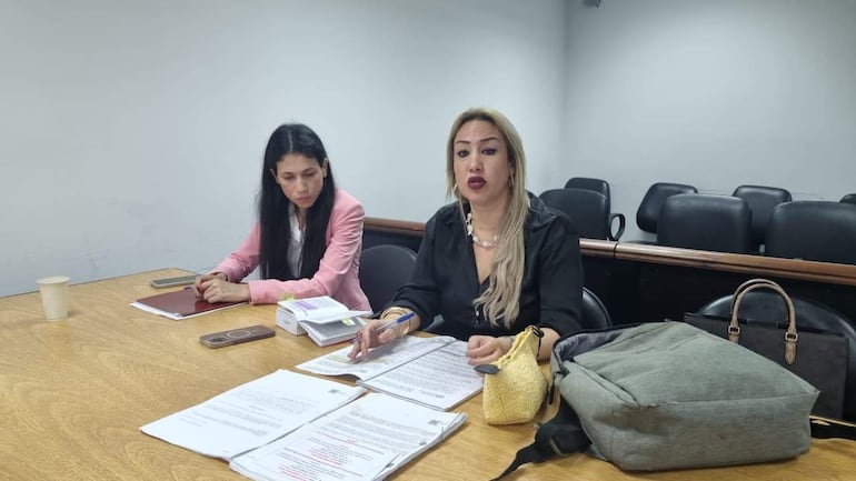 Mujer rubia en vestimenta negra habla con gestos, mientras mujer con cabello oscuro y prenda rosa escucha atentamente en mesa con documentos.