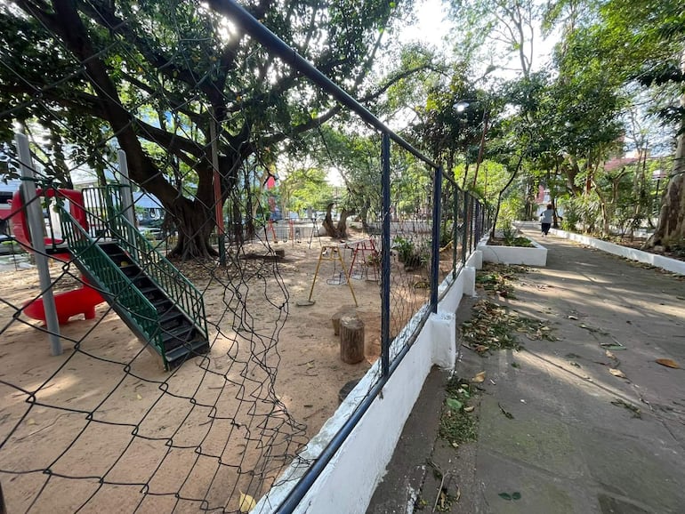 Área de juegos infantil cercada en plaza con densa vegetación y luz del sol entrando entre árboles.