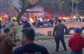Varias personas con chaquetas amarillas combaten un incendio en un área residencial, rodeados de humo y llamas.