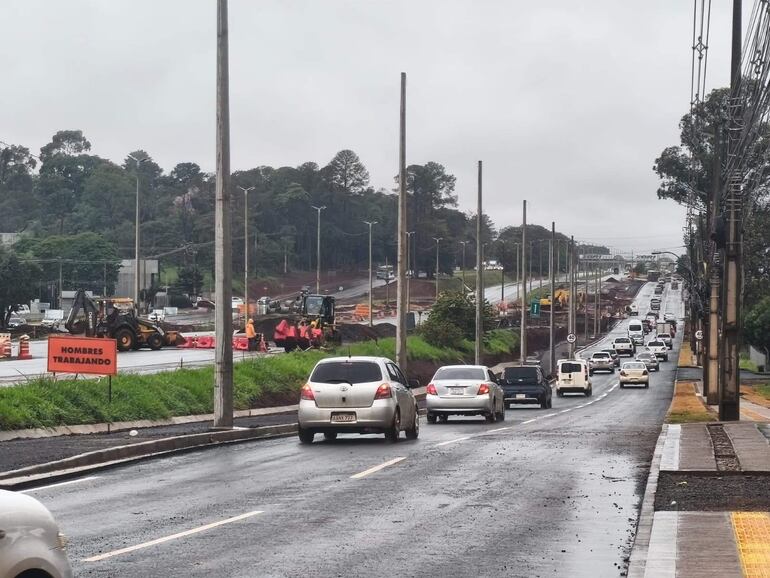 Trabajadores con chalecos naranjas en la carretera, rodeados de maquinaria pesada y vehículos en movimiento bajo un cielo nublado.