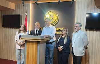 Hombre en el centro habla en conferencia con camisa clara, rodeado por mujeres y hombres escuchando atentamente en entorno institucional.