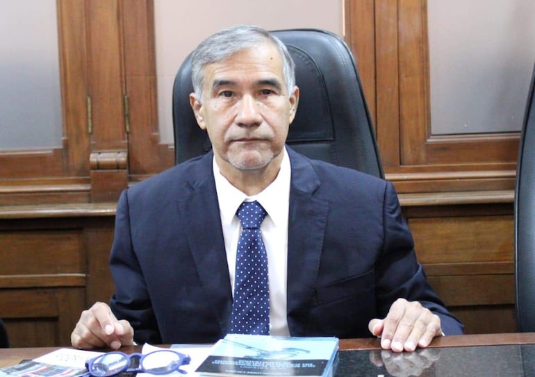 Hombre canoso en traje oscuro y corbata a rayas sentado en oficina con documentos sobre la mesa.