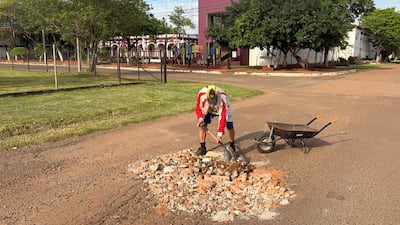 Hombre con camiseta clara y chaqueta roja, usando pala y carretilla vacía, en un entorno residencial con parque infantil.