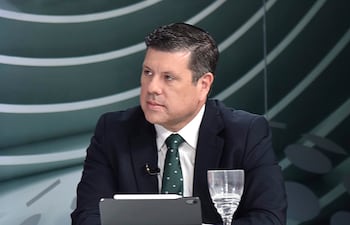 Javier Giménez, vestido con traje oscuro y corbata verde, escucha atentamente en un set de televisión con tablet y vaso de agua.