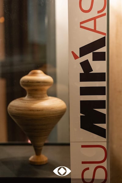 Un jarrón de madera en vitrina con diseño espiral, y cartel que dice 'SU MIRÁS' con letras rojas y negras.