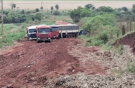 Tres camiones de carga en camino de tierra rojiza, rodeados de vegetación baja y campos de cultivo.
