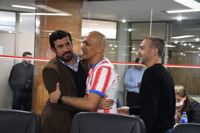 Juan José Arnold abraza a un hombre con camiseta rayada blanco y rojo, mientras otro observa en un ambiente de oficina.
