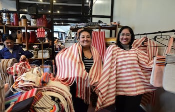 Mujeres con ponchos de rayas blancas y rojas sonríen mientras sostienen prendas en un ambiente de venta de artesanías.