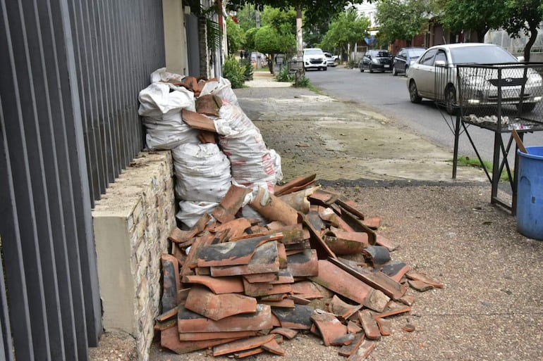 Pilas de tejas rotas y bolsas blancas amontonadas en la acera de una calle residencial sin presencia de personas.