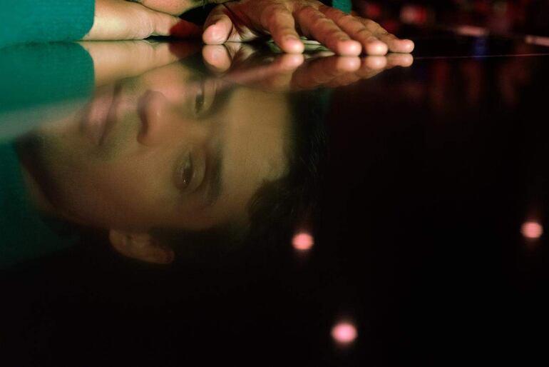 El David Aguilar, vestido casual, parece relajado con una mano sobre la mesa y rostro introspectivo en un ambiente de luz tenue.