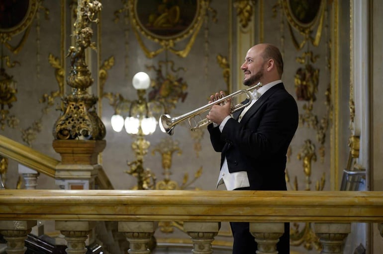 Hombre en traje negro y pajarita blanca, tocando trompeta con expresión concentrada en un ambiente lujoso.