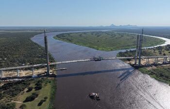 Vista aérea de un puente colgante en construcción sobre un río marrón, rodeado de vegetación densa y montañas al fondo.