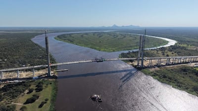 Vista aérea de un puente colgante en construcción sobre un río marrón, rodeado de vegetación densa y montañas al fondo.
