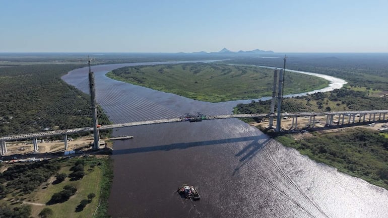 Vista aérea de un puente colgante en construcción sobre un río marrón, rodeado de vegetación densa y montañas al fondo.