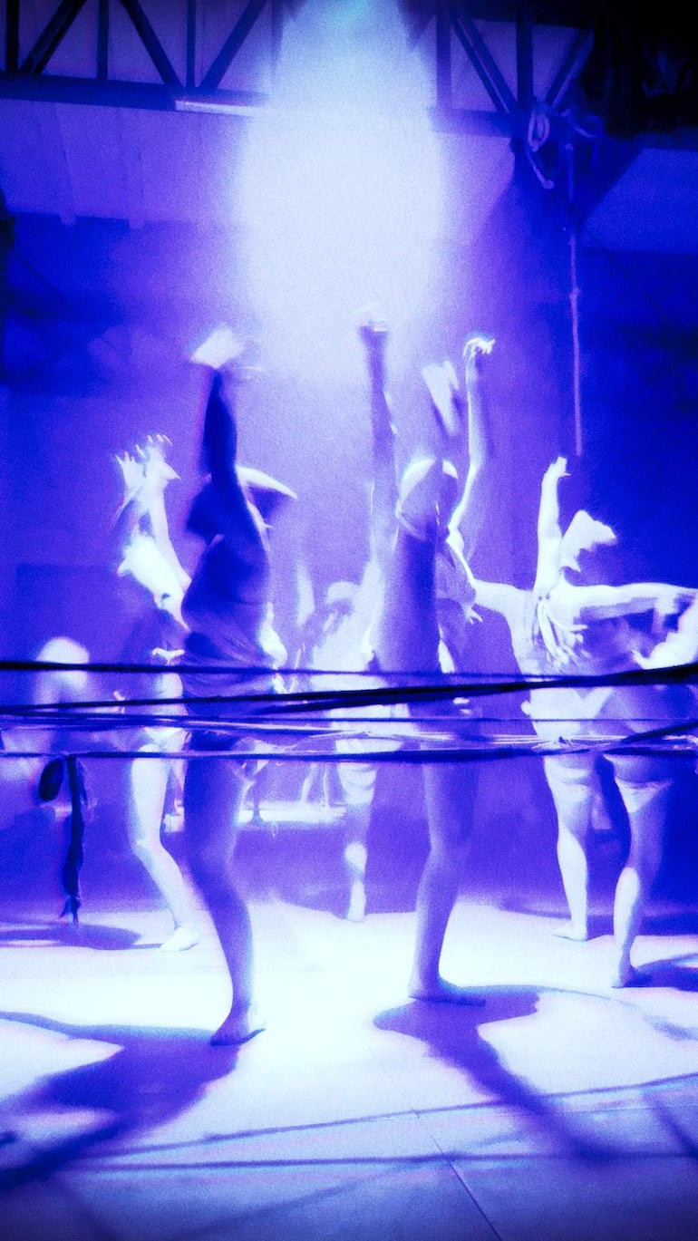 Cinco bailarines en el escenario, con brazos en alto y luces azules creando una atmósfera dramática y artística.