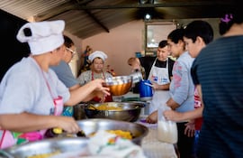 Mujer con delantal rojo y gorro supervisa mientras otros sostienen recipientes y preparan alimentos en una cocina.