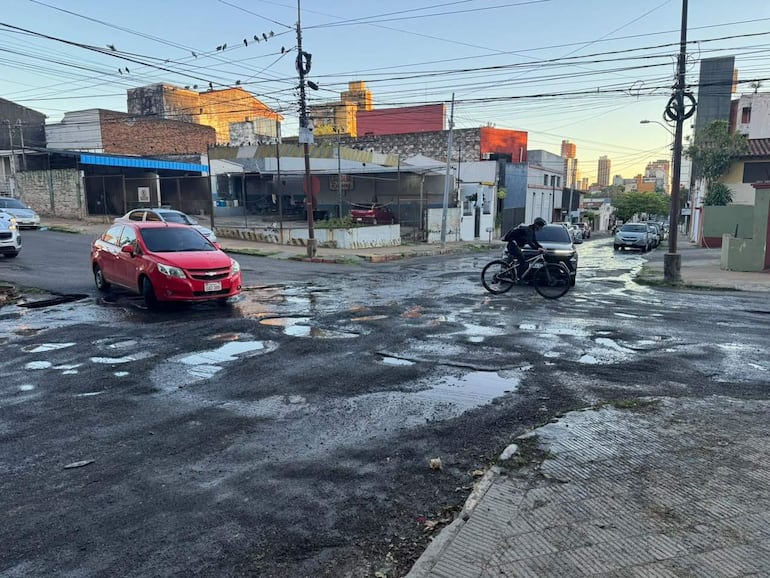Automóvil rojo y ciclista en calle con baches y agua acumulada, entorno urbano claro y edificios sencillos al fondo.