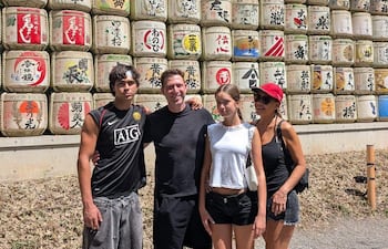 Cuatro jóvenes sonrientes con ropa casual posan frente a barricas de sake decorativas en un entorno al aire libre.