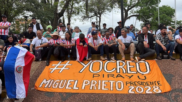 Grupo de personas con camisetas a rayas rojas y blancas, un hombre cubierto con una bandera paraguaya, observa un gran cartel de campaña.