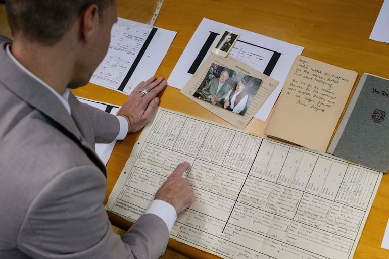 Esta fotografía, tomada el 2 de junio de 2026, muestra al profesor alemán Felix Puelm observando los documentos del «certificado de ascendencia» de su abuelo Paul (centro), copias impresas de carnés de afiliación al Partido Nacionalsocialista Obrero Alemán (NSDAP) de varios familiares, una fotografía de sus abuelos Paul y Brigitte, una pequeña imagen de su abuelo, una carta personal escrita por Brigitte después de la guerra y una copia impresa del carné de afiliación de Brigitte al NSDAP (arriba, centro). 