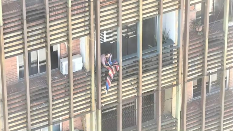 Persona asomada por el balcón de un edificio, sosteniendo una bandera roja, azul y blanca en un ambiente de humo.